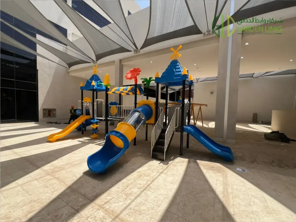 Playground equipment installation in Makkah with blue and yellow tube slide, yellow slide, blue slide, castle roof towers, white safety railings, and shade sail coverage