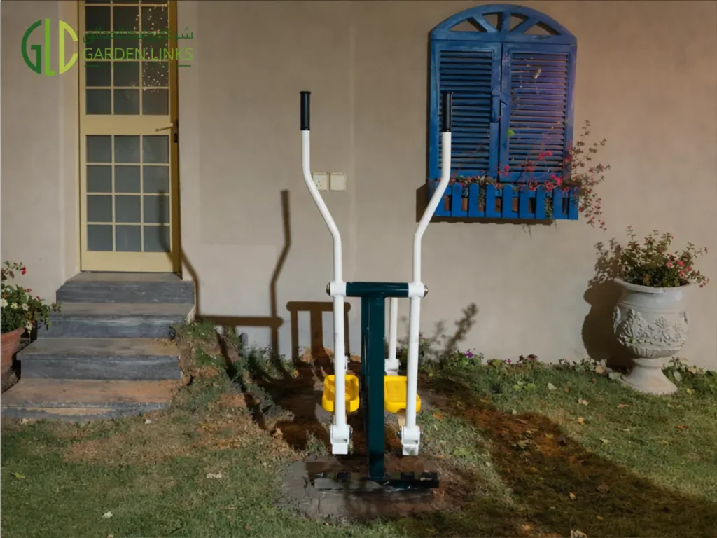 White and green outdoor elliptical cross-trainer fitness equipment installed beside a blue window in a Jeddah villa garden
