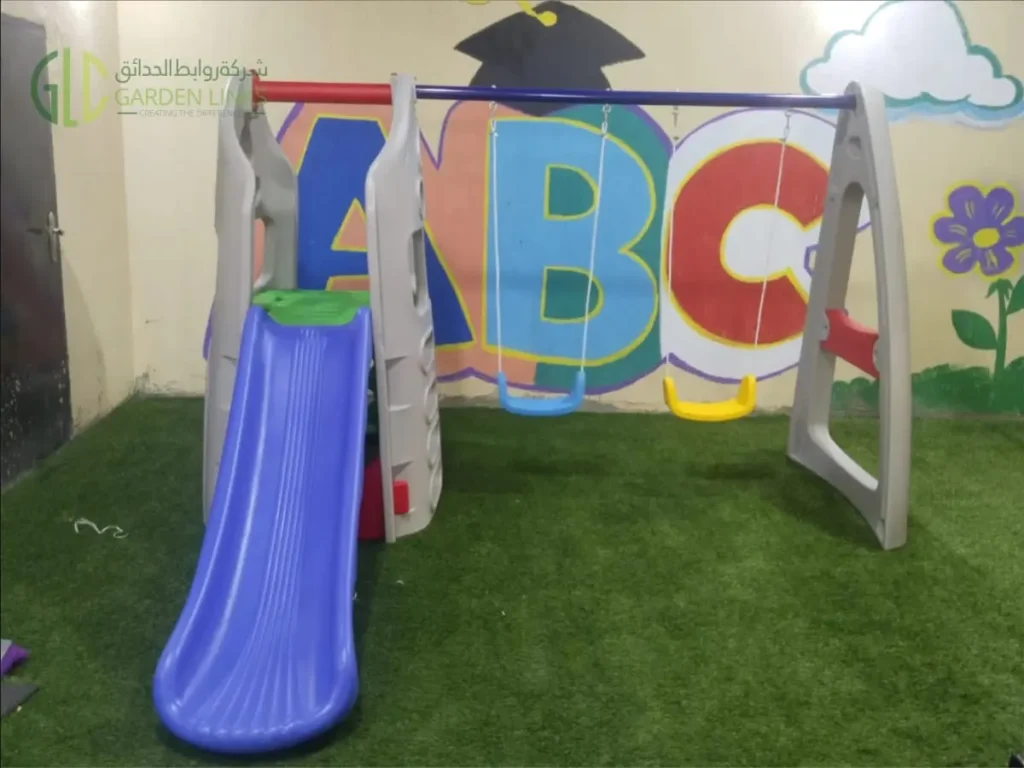 Children’s slide and two-seat swing set installed with artificial grass in an indoor school play area in Riyadh.