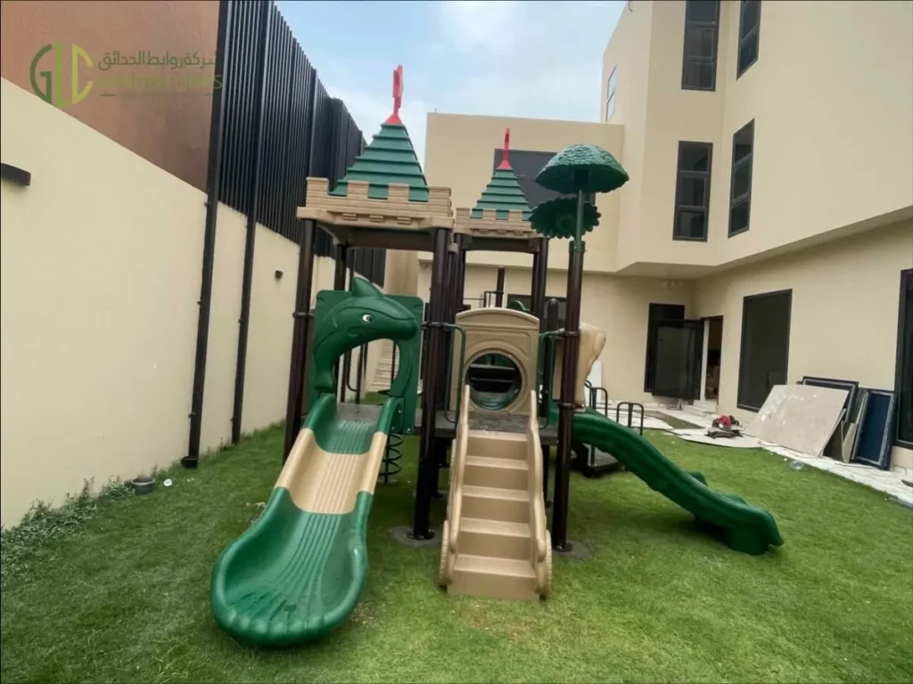 Outdoor school playground equipment in Riyadh with green double slide, spiral slide, access stairs, themed play towers, safety rails, and artificial grass.