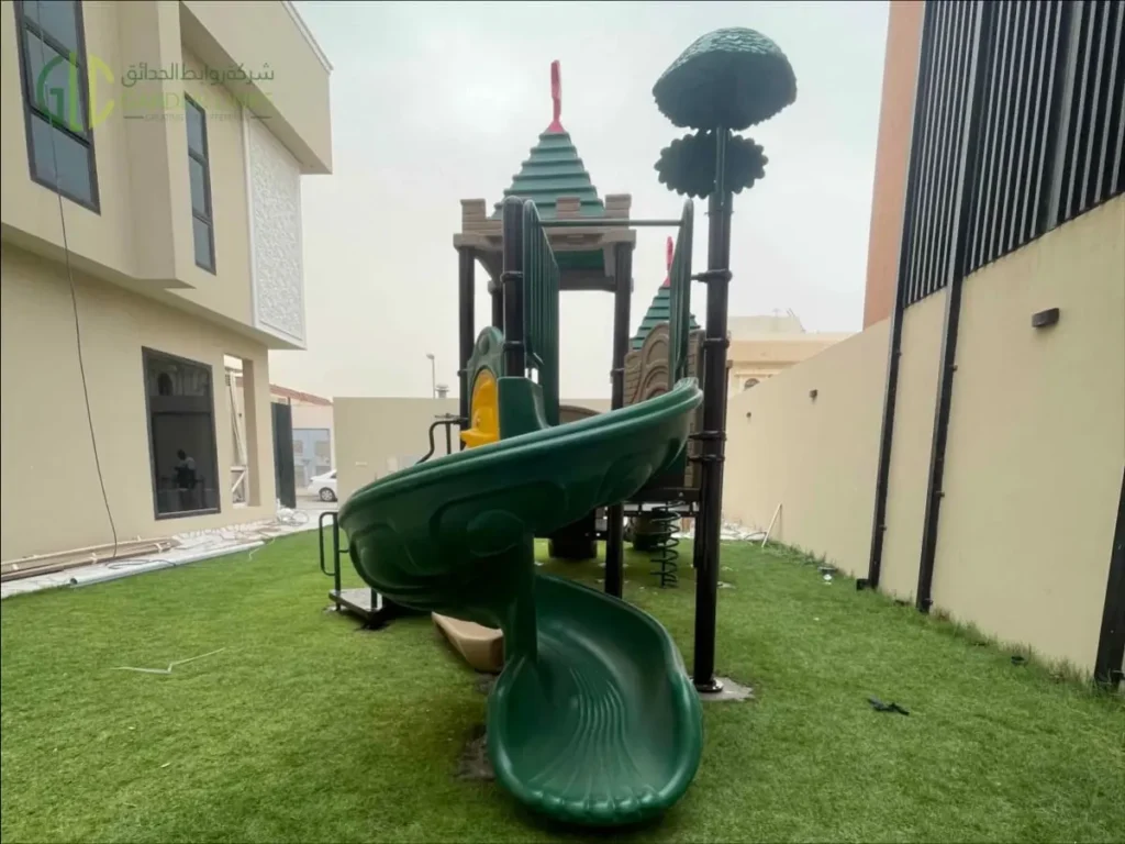 Outdoor school playground equipment in Riyadh with green double slide, spiral slide, access stairs, themed play towers, safety rails, and artificial grass.