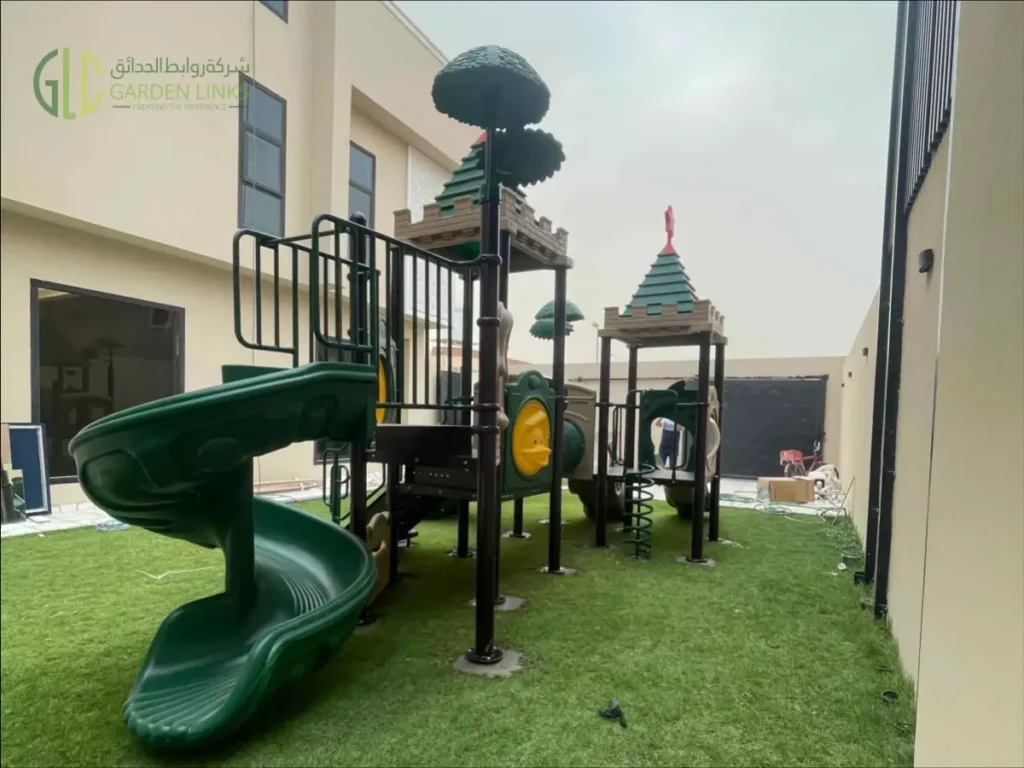 Outdoor school playground equipment in Riyadh with green double slide, spiral slide, access stairs, themed play towers, safety rails, and artificial grass.
