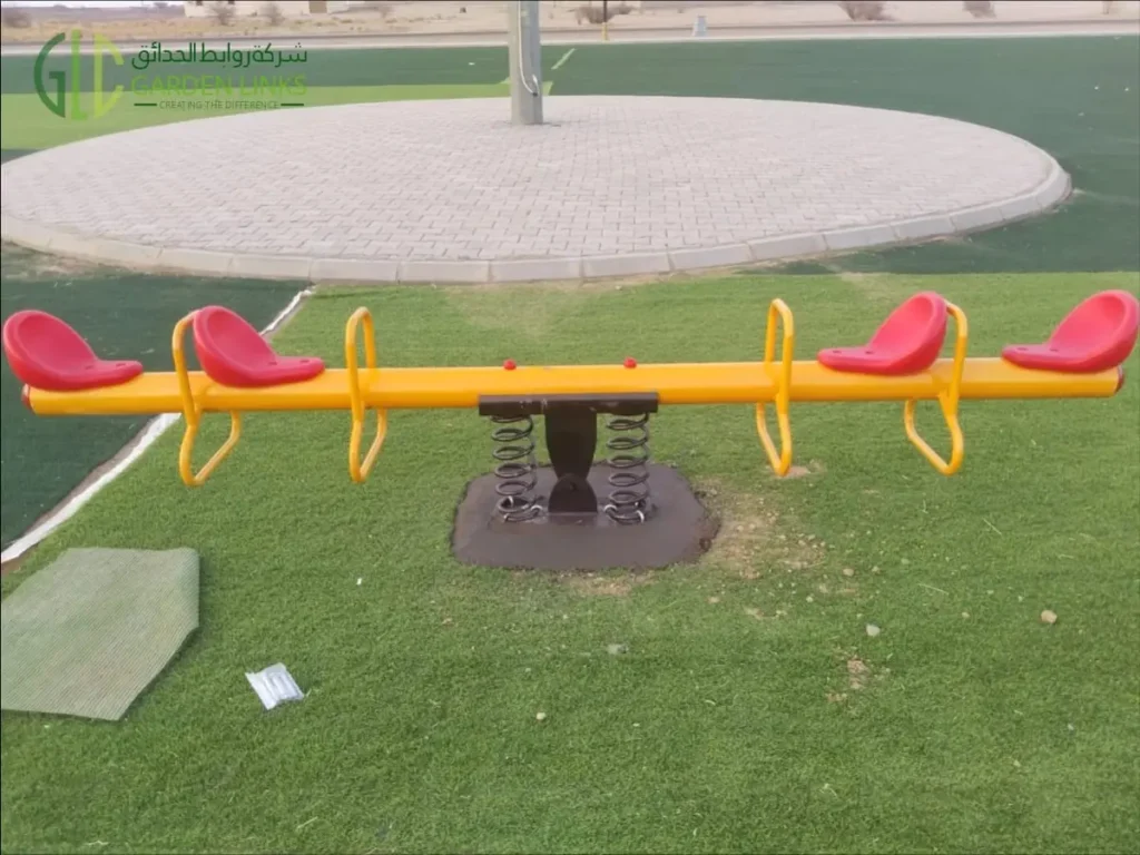 Four-seat spring seesaw installed at a public park playground in Madinah with red seats, yellow frame and spring support.