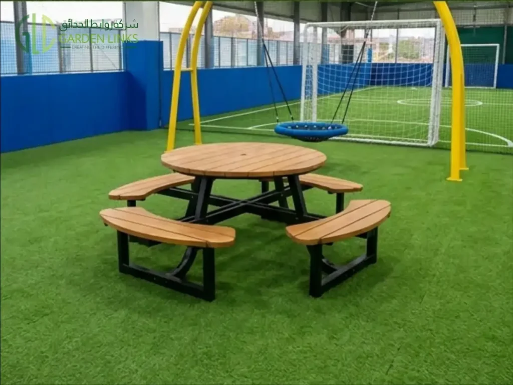 Round picnic table with curved benches in front of a blue basket swing on a yellow frame at Beech Hall School Riyadh.