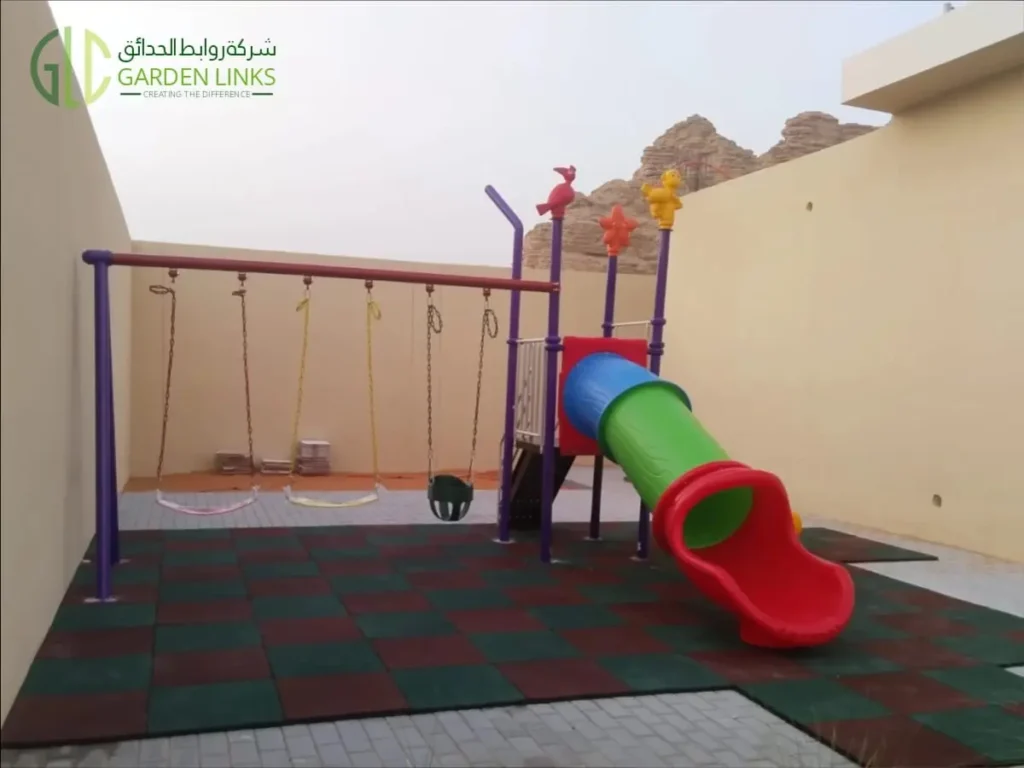 Double and tube slide home playset in Riyadh, viewed from the tube slide exit, with three swings and red and green safety flooring.