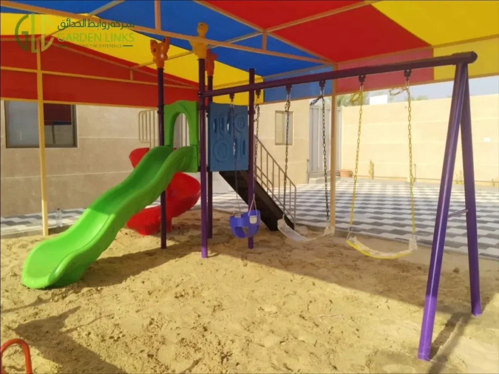 Shaded playset and seesaw installation in Banban showing three swings, a green straight slide and access stairs beneath a colourful canopy.